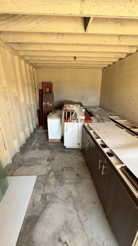Interior of a storage container with white walls, exposed beam ceiling, concrete floor, white appliances, and cardboard boxes stored inside.