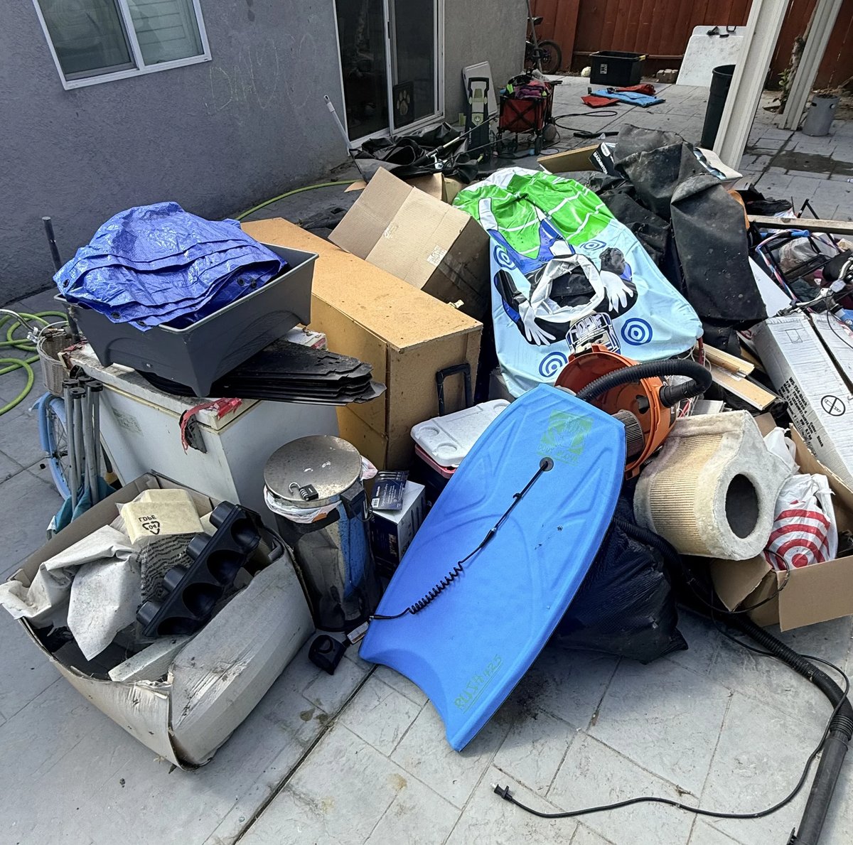 Pile of assorted junk and household items including boogie boards, cardboard boxes, and miscellaneous debris scattered on a driveway.