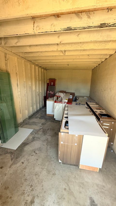 Interior of a storage container with wooden shelving, stacked white panels, boxes, and miscellaneous items on a concrete floor
