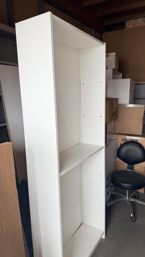 Empty white shelving unit with three compartments in a storage area surrounded by boxes and office furniture
