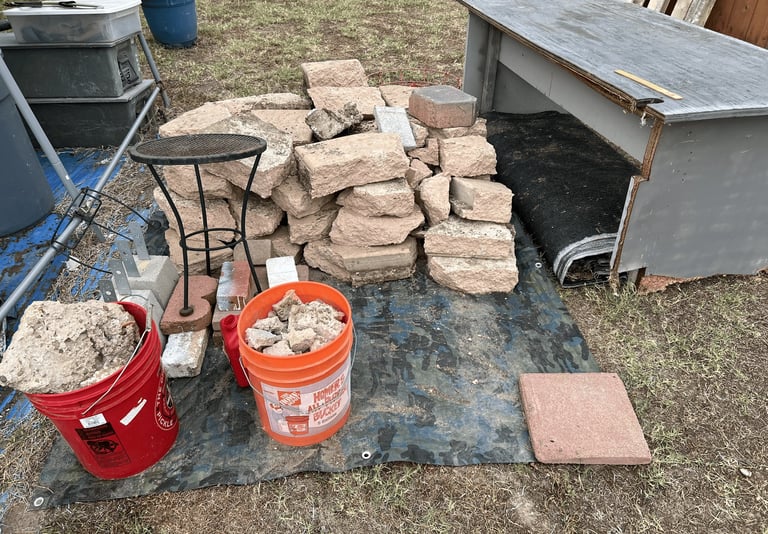 Construction materials including stacked bricks, red buckets, and pavers arranged on ground with metal containers and tools nearby