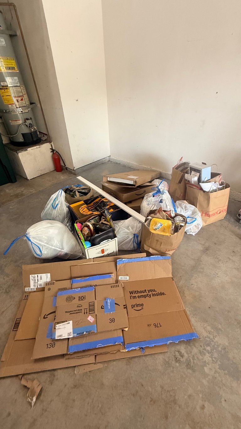 Cluttered garage floor with scattered cardboard boxes, plastic bags, tools in containers, and miscellaneous items awaiting organization or removal.
